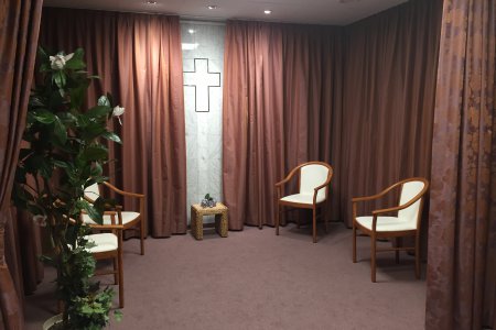 Ein Raum mit Teppich und warmen, gold-braunen bodenlangen Vorhängen ist zu sehen. Stühle, eine Pflanze und ein Kreuz an der Wand.
