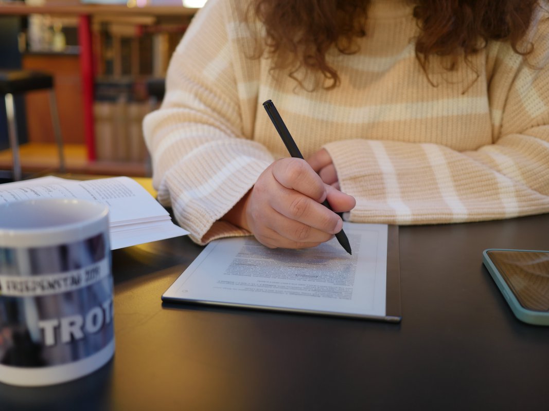 Jemand schreibt an einem Tablet, neben ihr ein Buch: Ein warmes Platzerl für Hausübungen und Seminararbeiten mit gratis Stromanschluss, Kaffee, Tee und Keksen